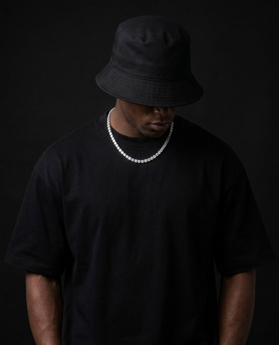 Person wearing a black bucket hat and black t-shirt with a tennis moissanite necklace on a dark background