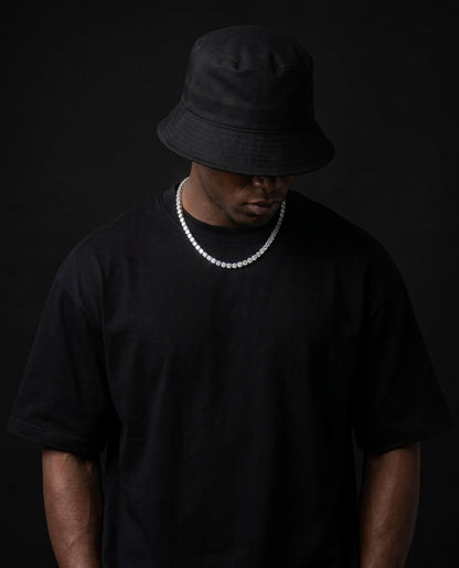 Person wearing a black bucket hat and black t-shirt with a tennis moissanite necklace on a dark background